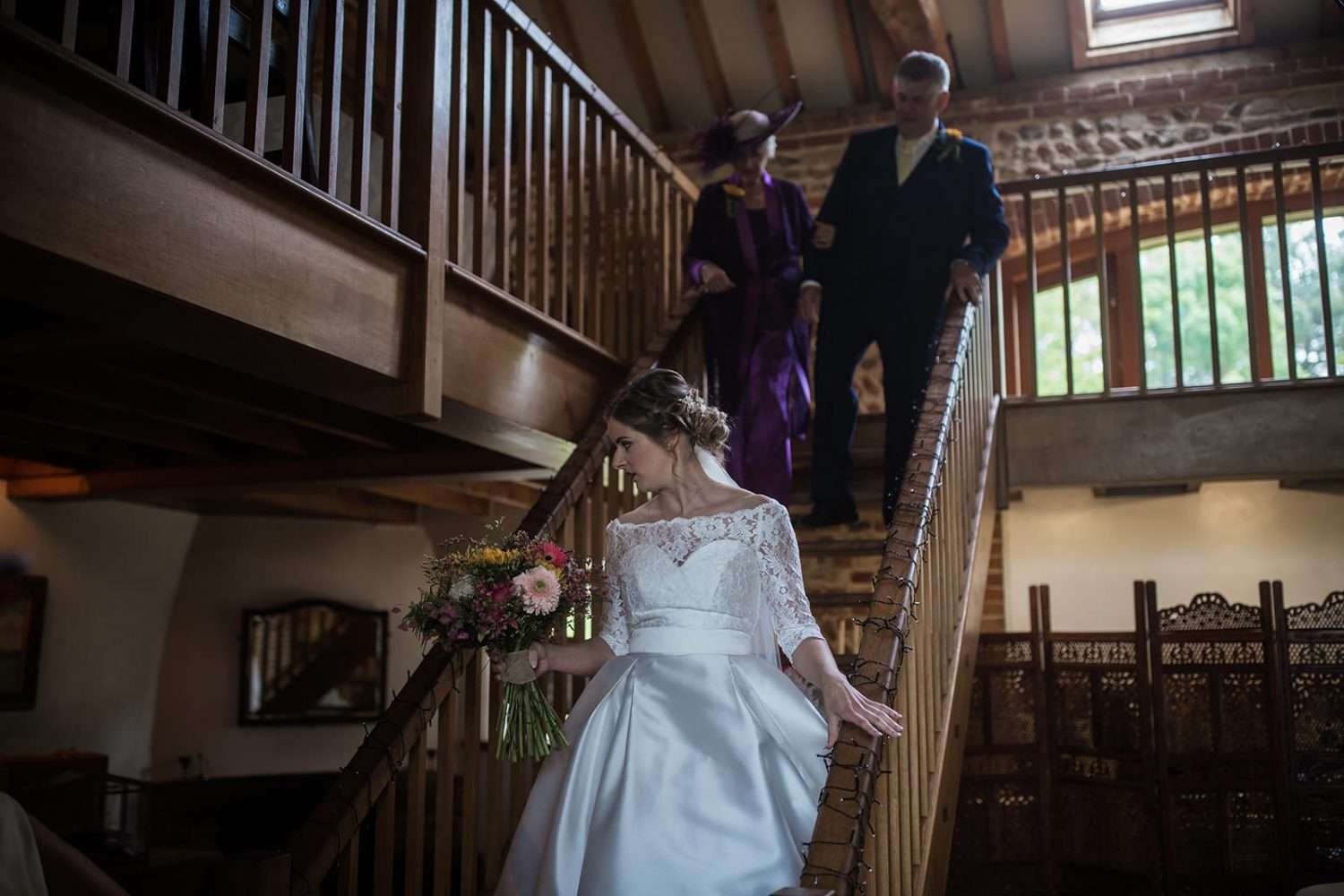 bride walks to her wedding ceremony at chaucer barn, she is accompanied by her mum and dad as they come down the stairs