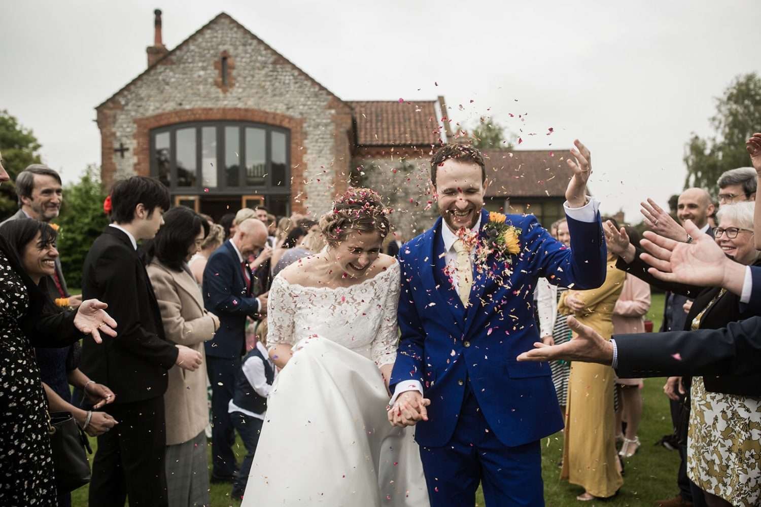 at a chaucer barn wedding we see the bride and groom laughing and walking through a funnel of their guests who are throwing confetti at them 