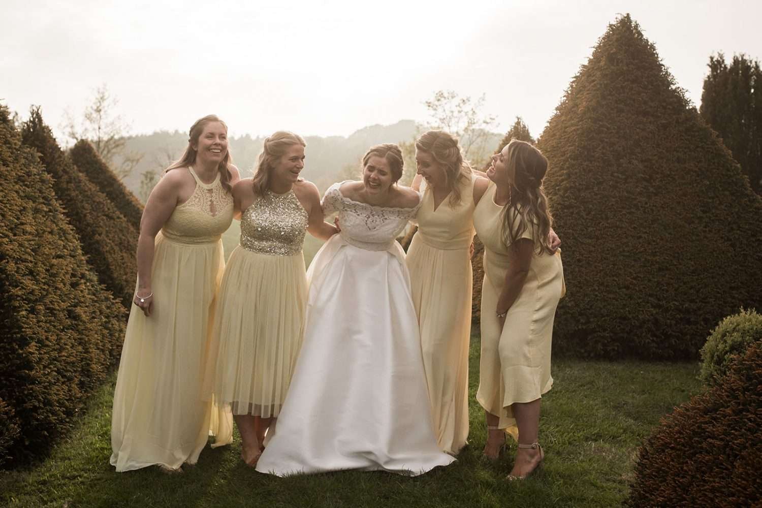 bride and her bridesmaids giggle during a photo at sunset, flanked by topiary bushes. 