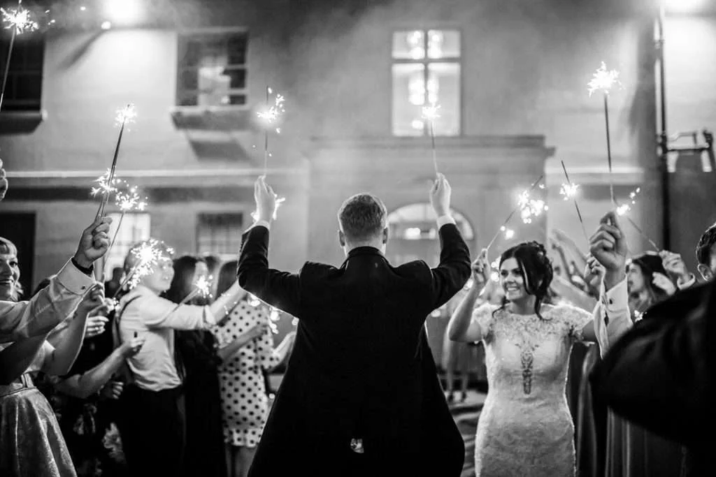 A Bride and Groom hold sparklers in their hands and kiss outside Lanwades Hall at their Suffolk wedding. They are surrounded by two lines of their family and guests also holding lit sparklers.