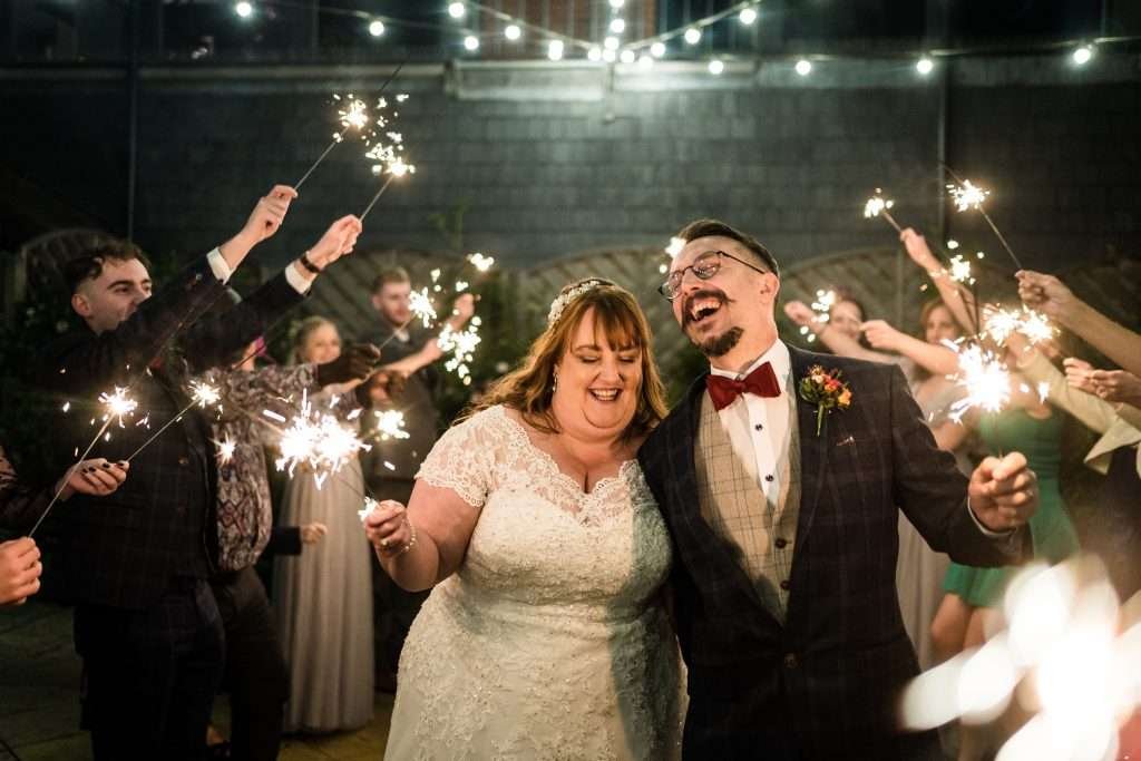 A Bride and Groom hold sparklers in their hands and laugh  at their Suffolk wedding. They are surrounded by two lines of their family and guests also holding lit sparklers.

