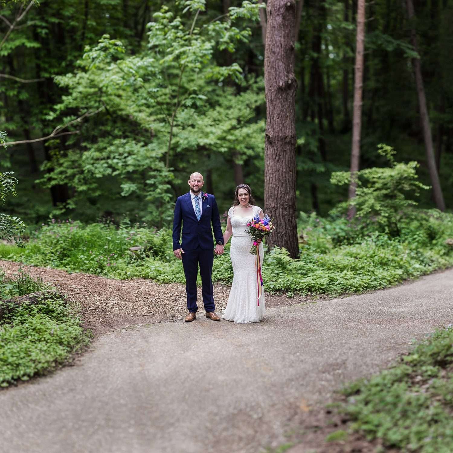 A Brenizer style portrait of a happy Bride and Groom holding hands after their wedding ceremony  in the beautiful Spring woodland landscape of Greenacres Colney Norfolk
