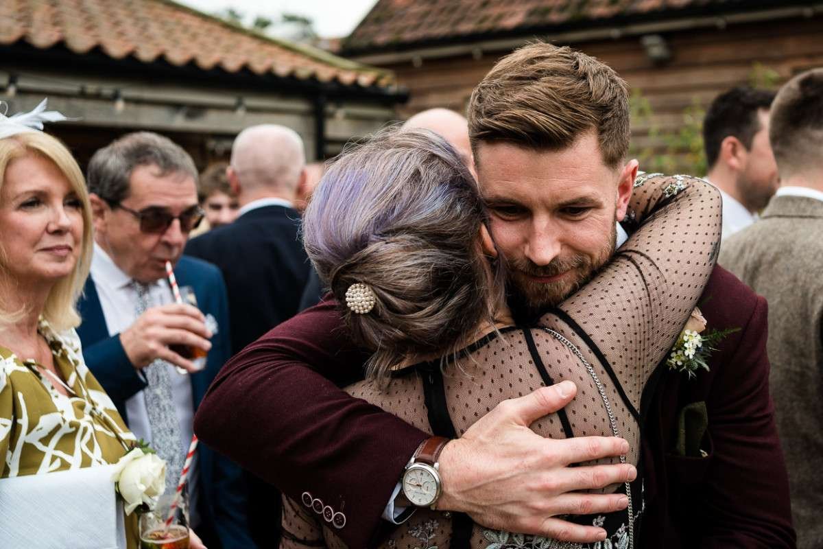 A Groom hugs a friend after his wedding ceremony whilst family members look on and smile. 
