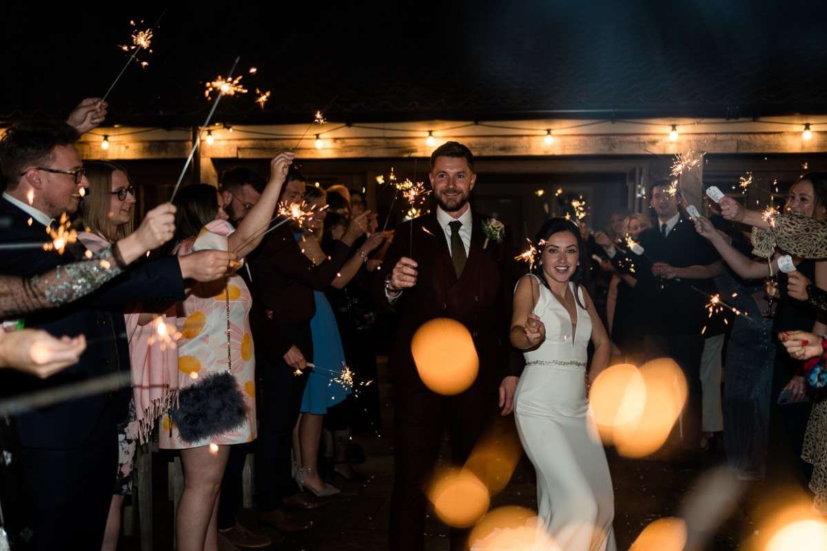 A Bride and Groom laugh whilst holding sparklers and walking between rows of their family and friends who also hold lit sparklers. It is dark and we are at the courtyard area wedding venue in Suffolk. 