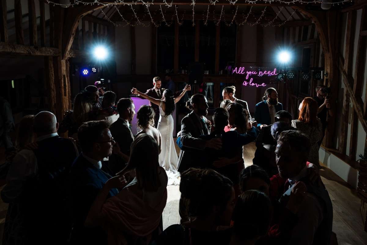 A couple hold each others outstretched arms and stare loving at each other towards the end of the first dance at their wedding at Easton grange In Suffolk. Around them their family and friends have joined the dancefloor and the lighting is moody and romantic. A neon sign to the right says 'All you need is love'.
