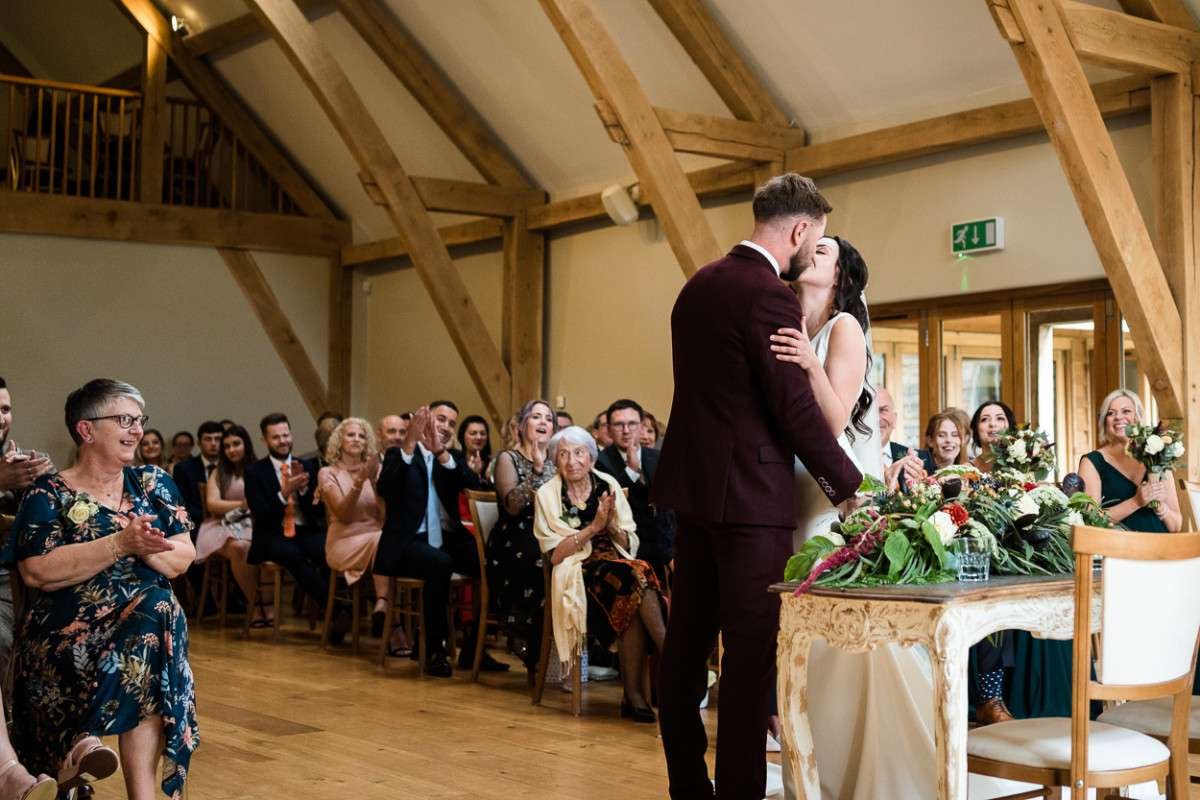 Bride and Groom kiss at the end of their ceremony on their wedding day at Easton Grange in Suffolk. Their family and friends applaud enthusiastically.