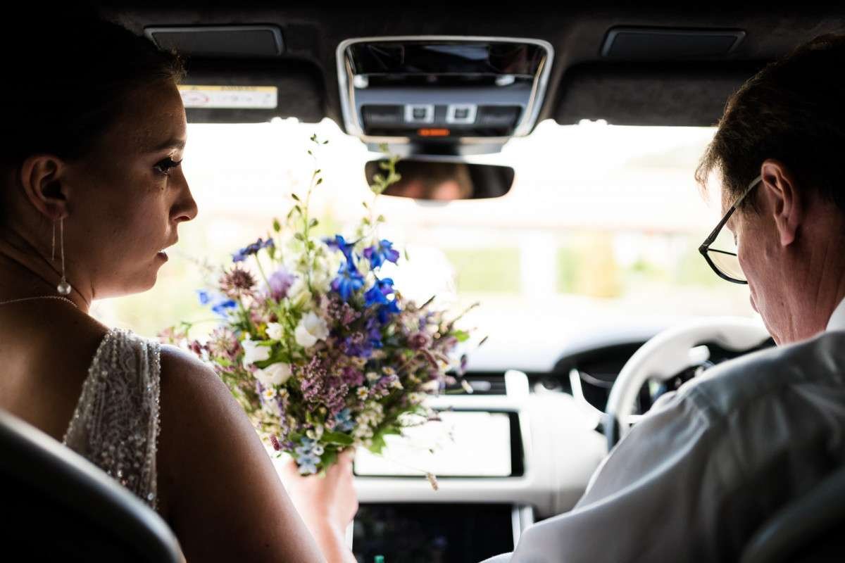 bride and her father en route to her wedding. She is holding a bouquet of flowers and looks to her dad who is driving the car. 
