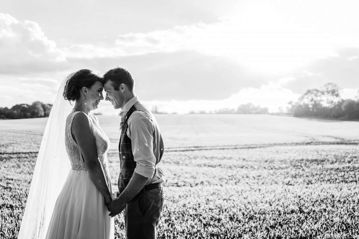 Newly married couple holding hands and pressing their foreheads together as they smile at their wedding in suffolk. They are lit by a setting sun in a filed of barley. 
