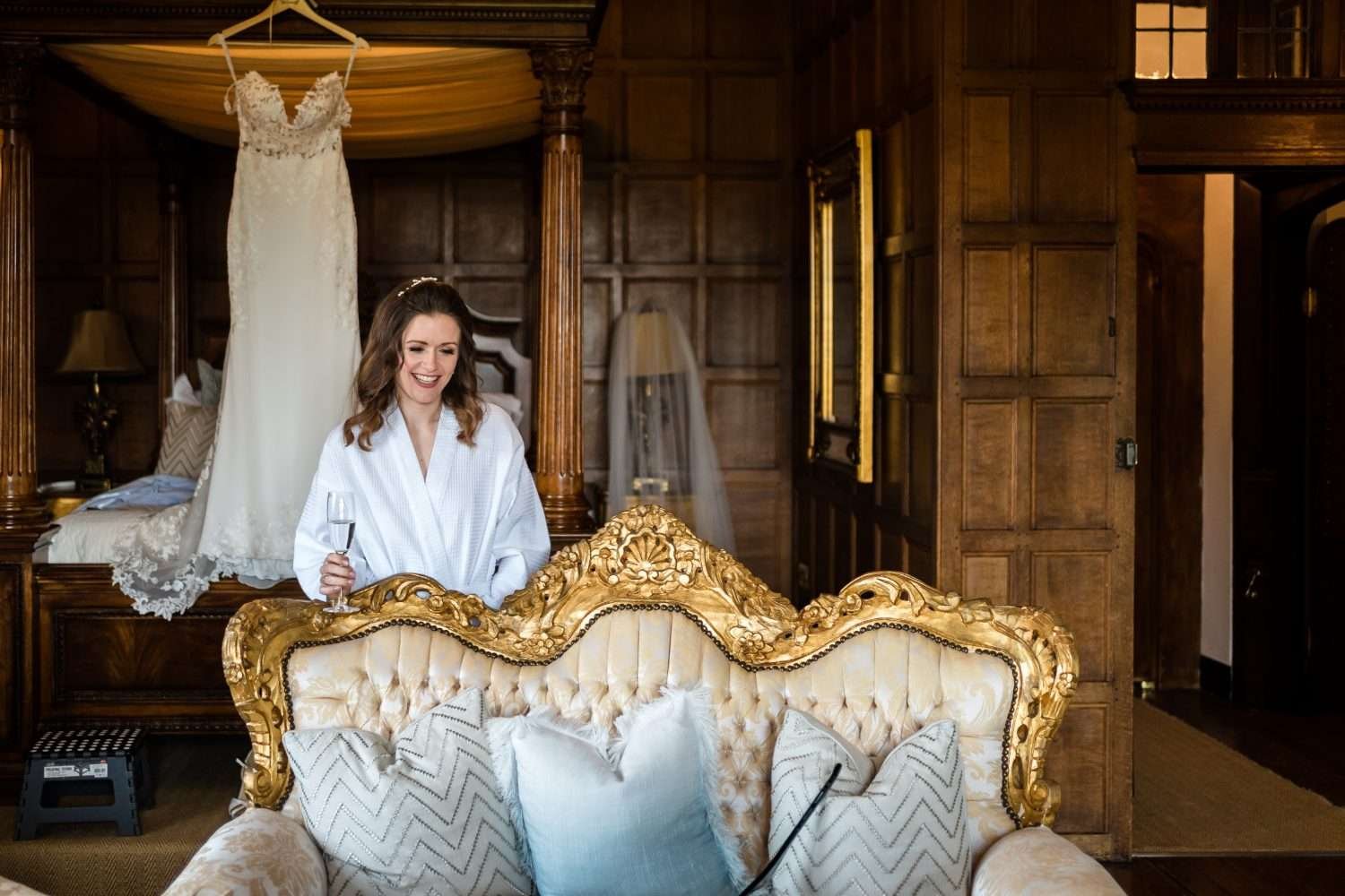 A bride is getting ready for her wedding but she is still in her dressing gown and drinking wine while wearing her tiara! She is standing behind a richly decorated sofa in a wood panelled elizabethan room and her wedding dress is hanging from a four poster bed behind her. 