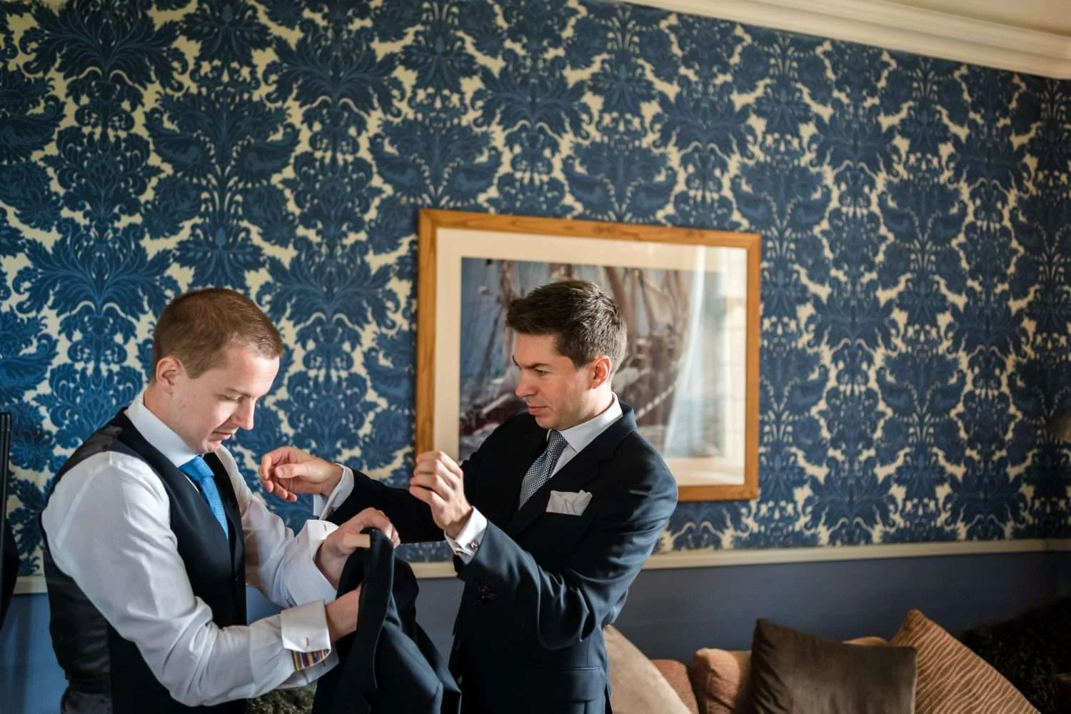 A groom and his grooms man getting ready for the wedding in a blue wallpapered room at hengrave hall in Suffolk UK 