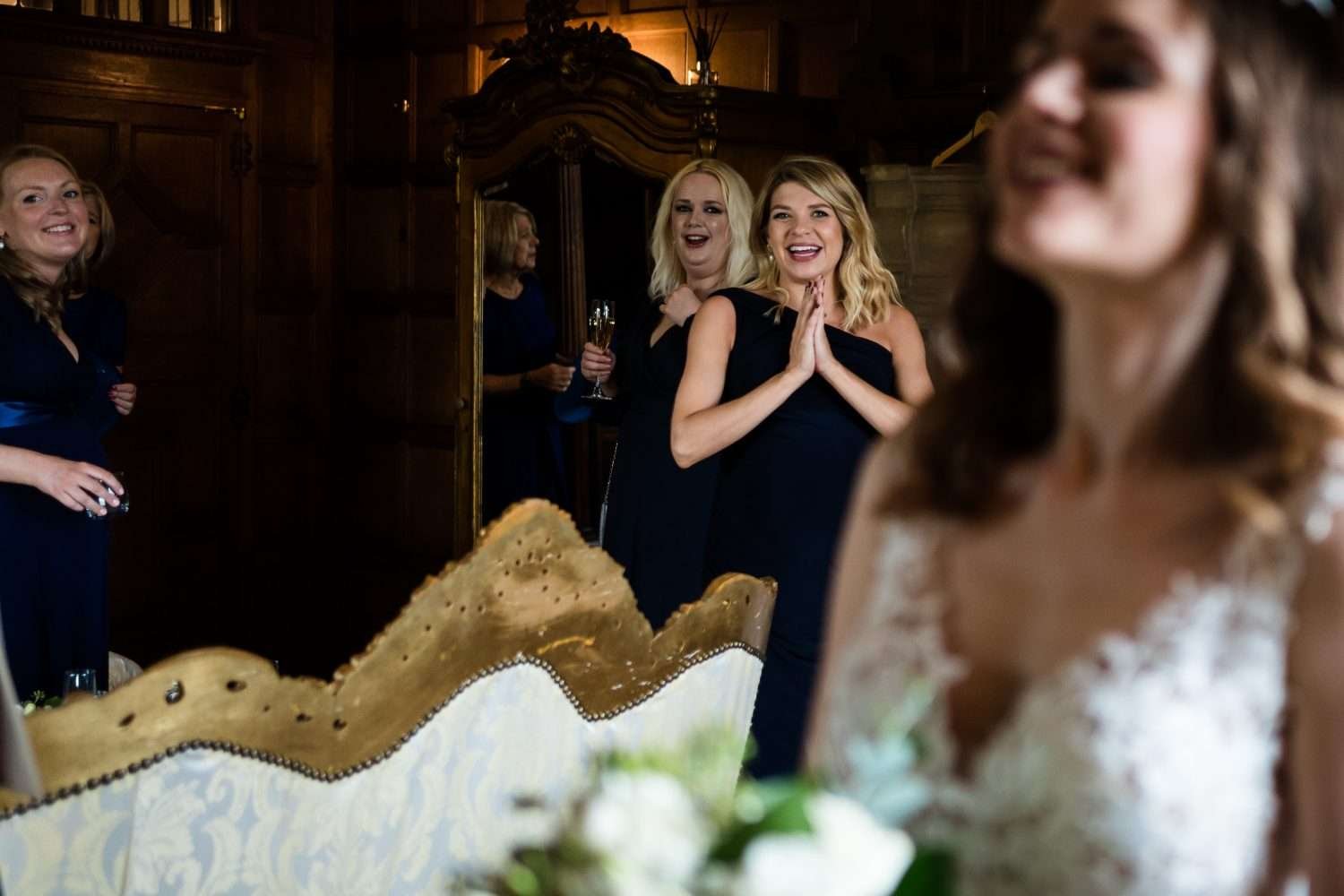 Bridesmaid smile excitedly to see the bride in her dress for the first time. The bride is visible smiling but out of focus in the foreground. 