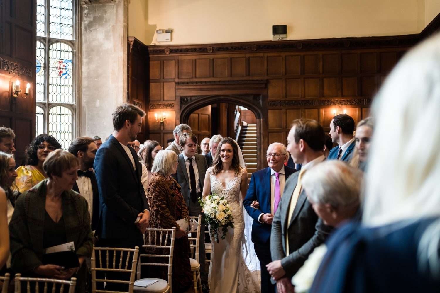 A Bride is escorted down the aisle by her father at her wedding. She is smiling at her groom (not shown) as she moves through the congregation of family and friends who are all smiling at her as she passes. 