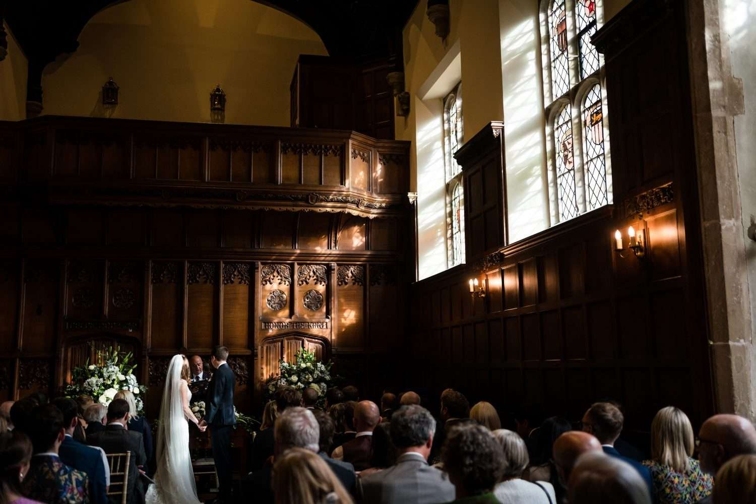 Sunlight streams from high elizabethan leaded windows on the right of a wood panelled room where a bride and groom are being married amid a crowd of seated family and friends. 