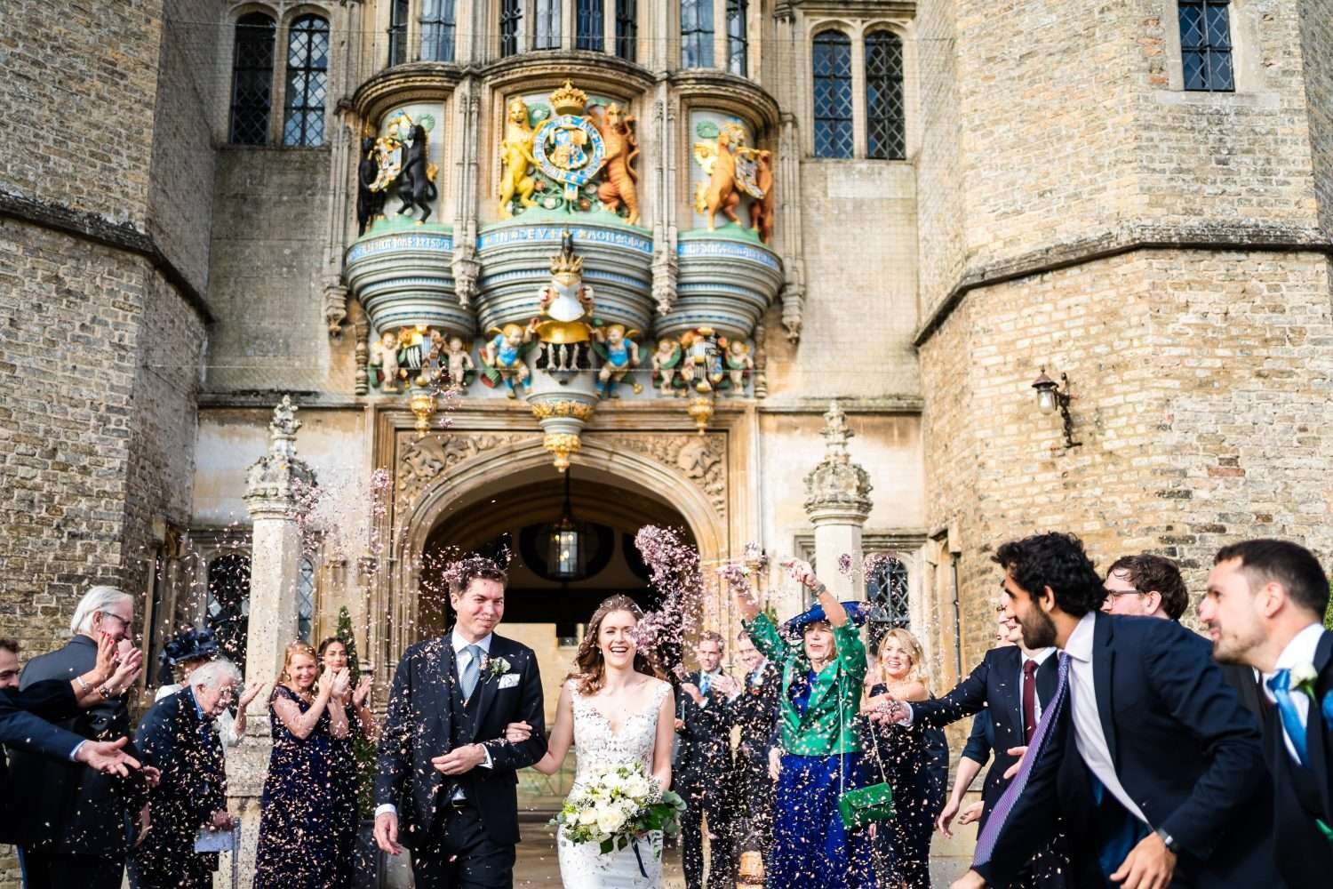 A couple are overwhelmed by confetti thrown by their guests at their wedding at hengrave hall. We see the front door and associated stonework of hengrave hall in the background. 