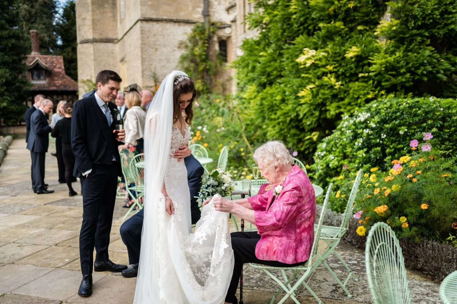 a bride shows her grandmother her wedding dress on the west terrace at hengrave hall in suffolk whilst her groom looks on 