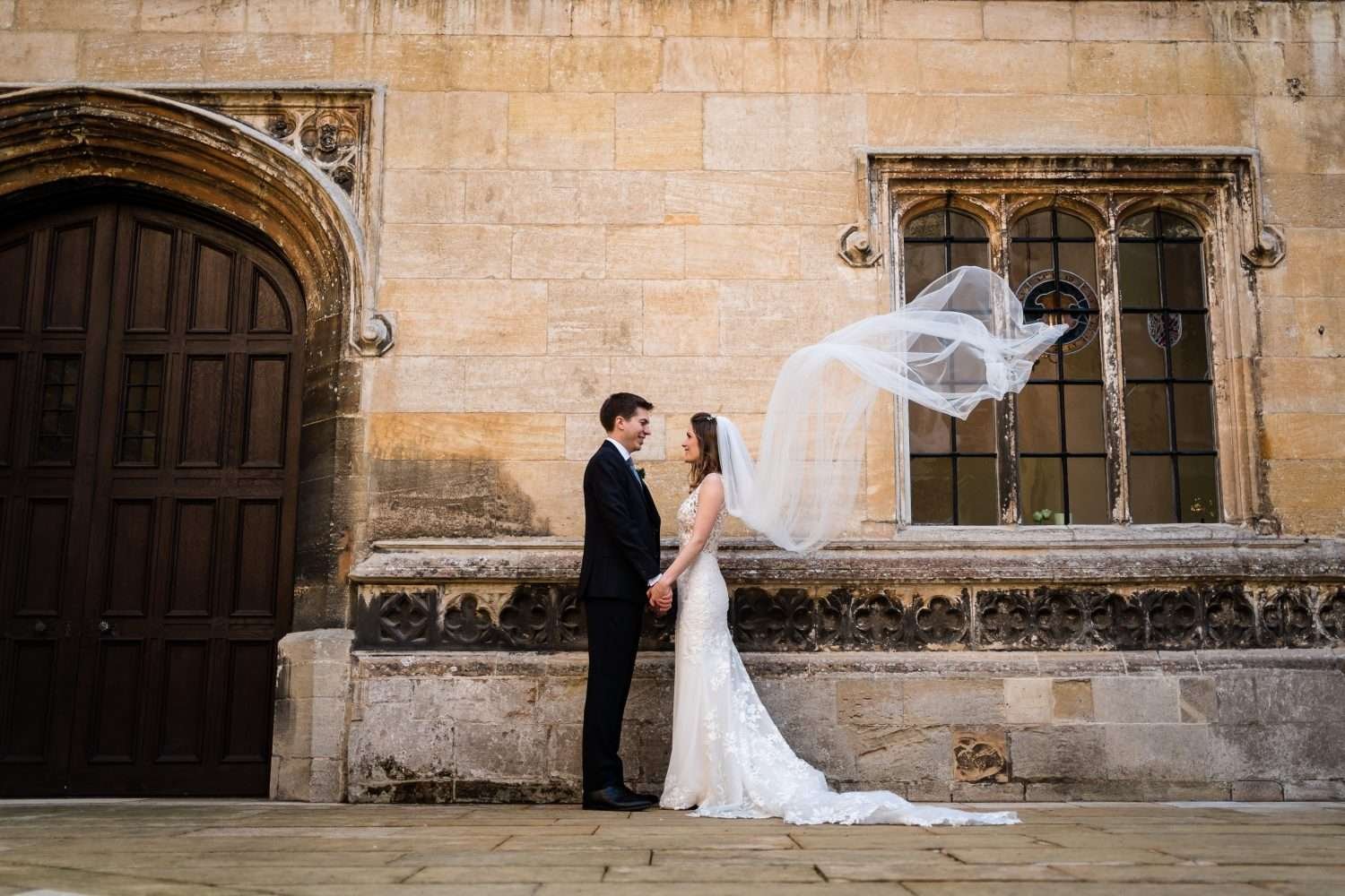 wedding portrait at hengrave hall in suffolk. The couple face each other while holding hands in the courtyard. The brides veil is caught by the wind and swirls behind her. 