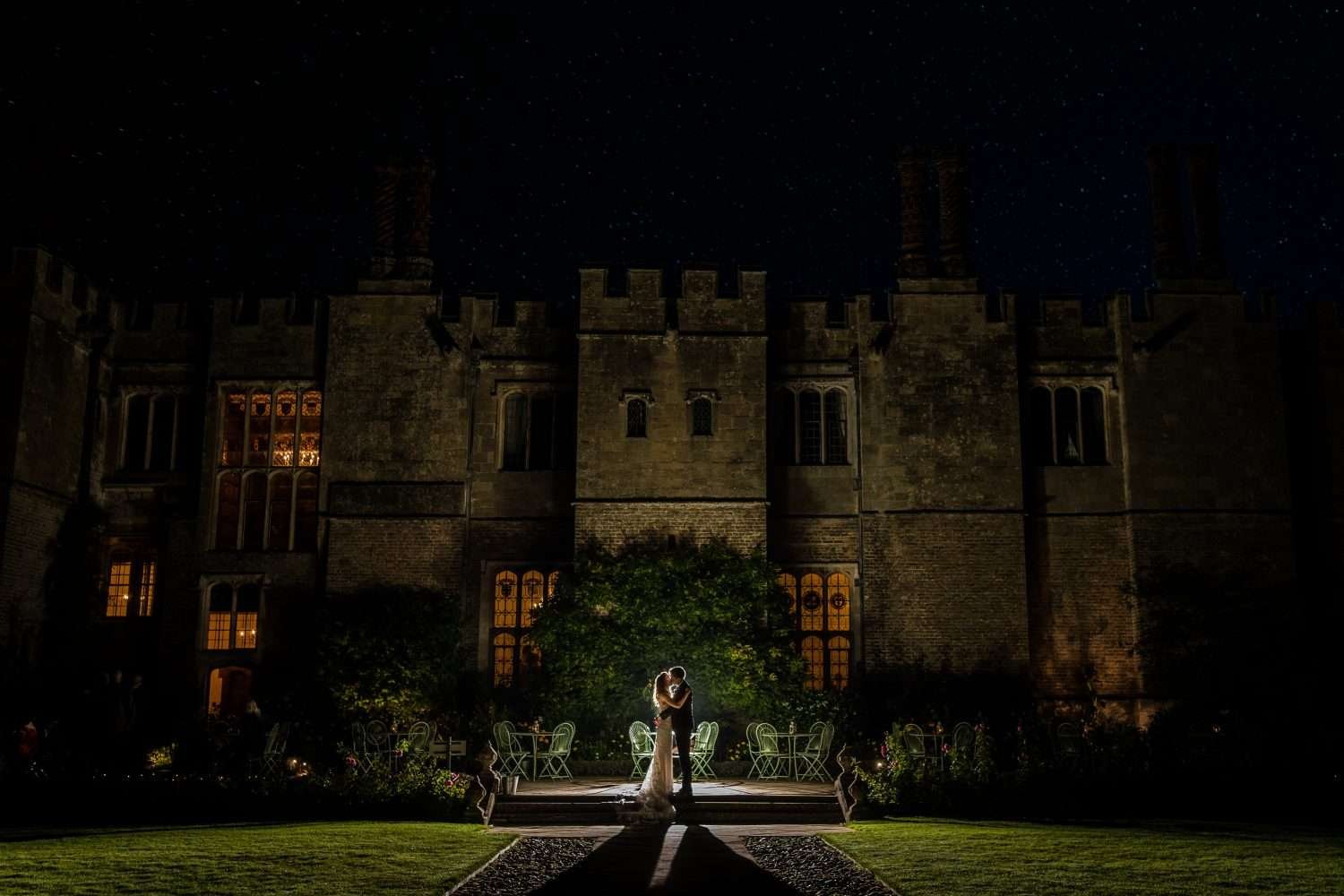 Hengrave Hall Suffolk - A bride and groom portrait taken at night on the west terrace at hengrave. The couple are chest to chest and kiss with backlit flash illuminating them. The building is also partly lit by the flash but also from the internal lights seen through the windows. The grass and climbing plants are radiant and sparkly. 