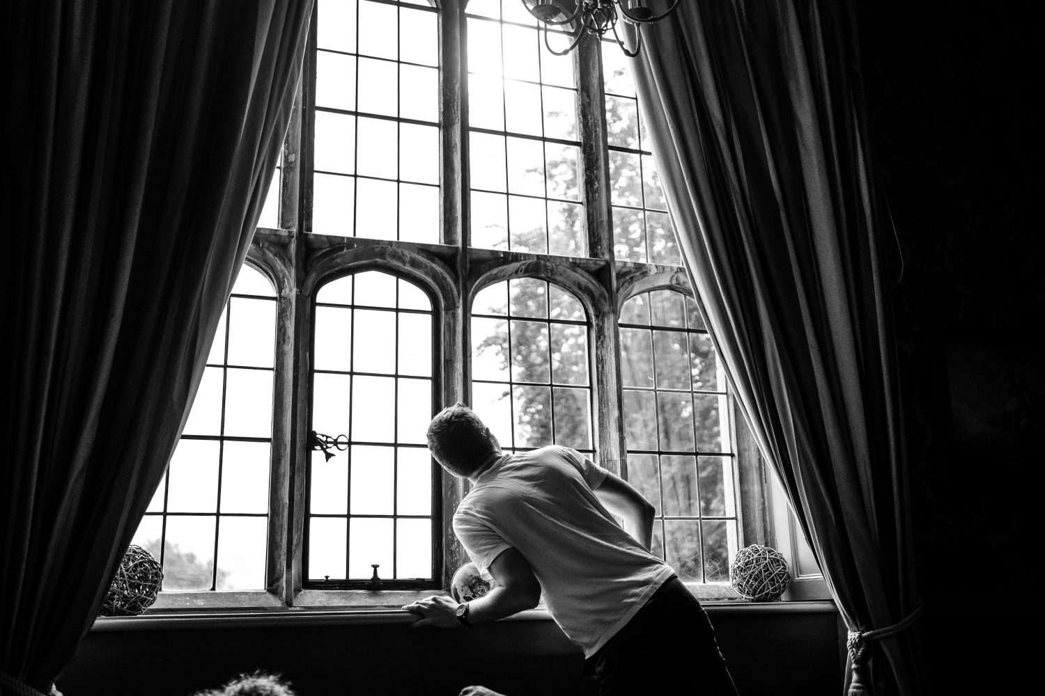 Black and white photo of a groom before he is ready for his wedding as he checks out the large elizabethan window of his room to see what the weather is doing. 
