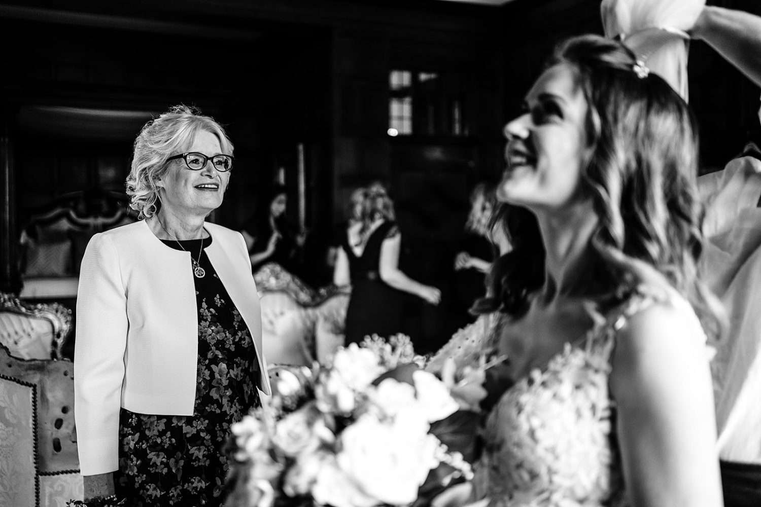A Brides Mum sees her Daughter in her wedding dress for the first time at Hengrave Hall. The Brides Mother is smiling in sharp focus whilst we see her Daughter laughing in the foreground and having her veil fitted. To the rear are Bridesmaids looking on. 
