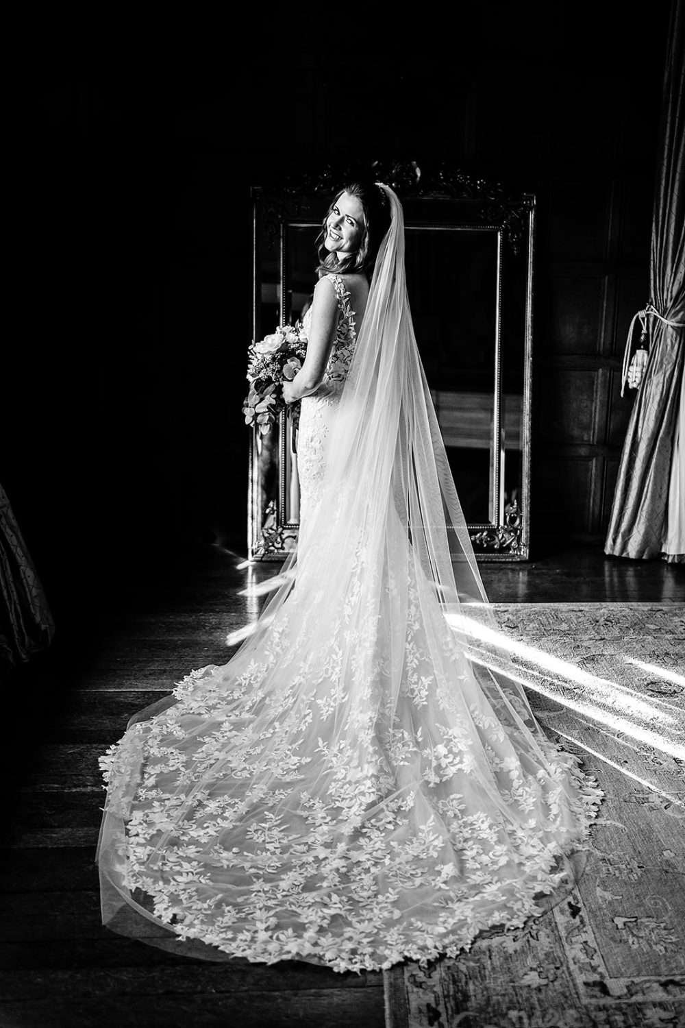 A Black and White photograph from a Hengrave Hall wedding. We see the Bride in the last moments of getting ready. She is in her wedding dress and veil whilst holding her bouquet. The Bride is looking over her shoulder and smiling to camera and her dress and face are lit by sunlit coming from the window to the photos right. 