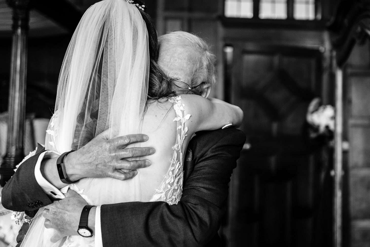 A Father of the Bride hugs his Daughter hard having seen her in her wedding dress for the first time. The Black and White photograph records an Elizabethan panelled room and we see some of the face of the elederly Father and just the back and white veil of his Daughter. 
