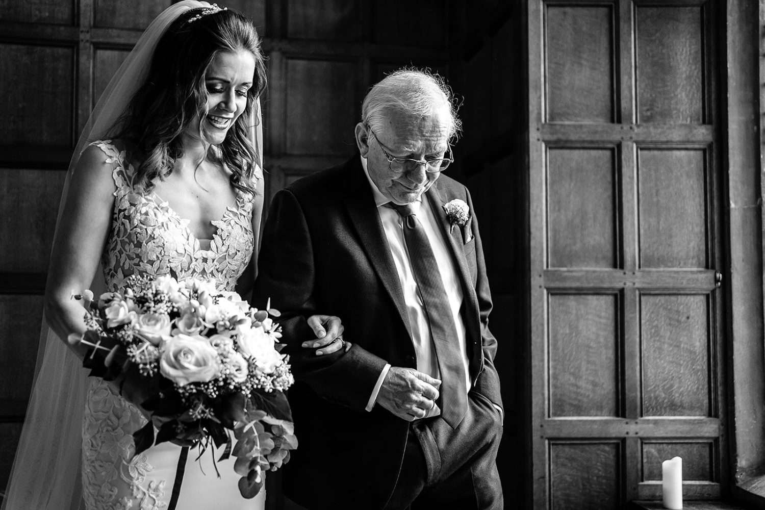 A Father escorts his Daughter at her Hengrave Hall wedding day on the way to the ceremony. This Black and White photo shows the elederly Father supporting his Daughter by taking her arm as they descend the stairs. The Bride is talking to her father and also holding her bouquet. 
