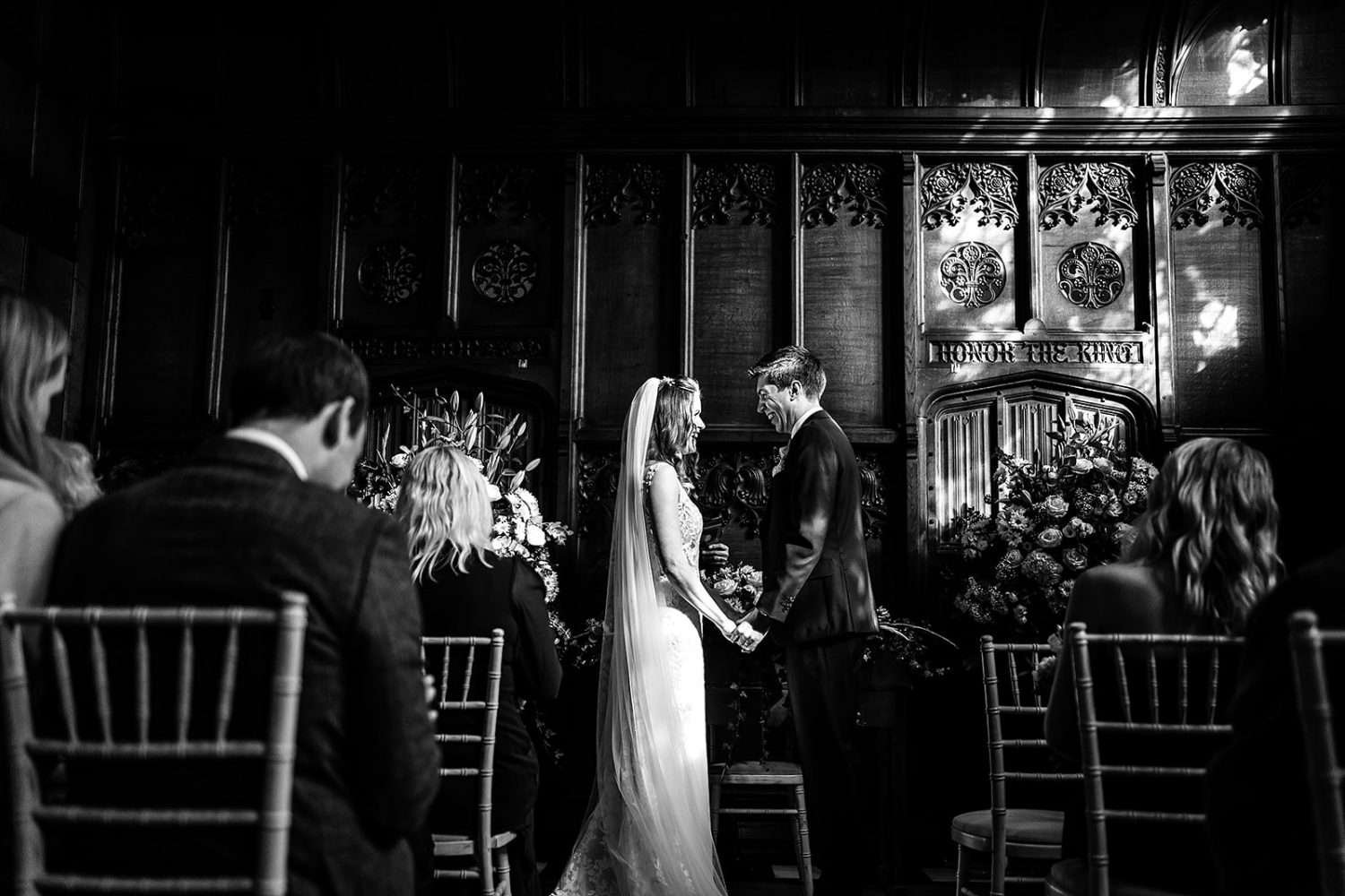 A black and white photo from a wedding in Suffolk at Hengrave hall. We see a Bride and Groom holding hands and smiling at one another just before the kiss. The Groom wears a black traditional tuxedo and the Bride is in a white lace full length dress with cathedral veil. The wood panelling of the great room is in the background and a floral arrangement is to the left of screen. 