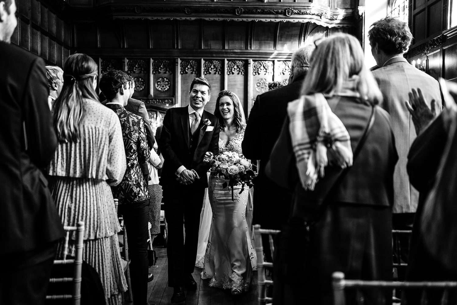 A black and white photo from a wedding in Suffolk at Hengrave hall. We see a Bride and Groom arm in arm and smiling as they leave their wedding ceremony. Their guests and family are clapping and smiling too. The groom is in morning suit and the brisde wears a lace dress with veil. 