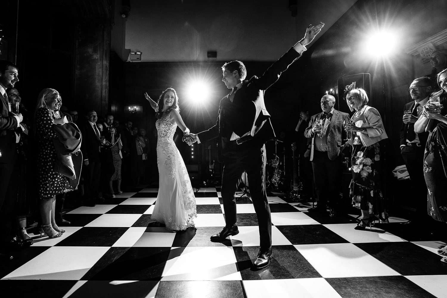 A bride and groom perfrom their first dance at their wedding. They are holding hands and in a dramatic pose. The couple are backlit by flash light and surrounded by their family and firnds who are smiling. The dance floor is covered in black and white tiles like a chess board.  