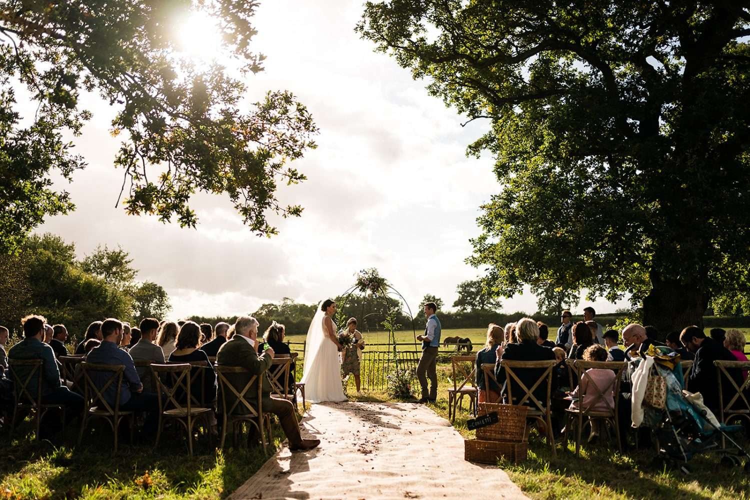 A bride and groom at their outdoor festival style wedding ceremony in Suffolk. We see the couple listening to their celebrant as their seated guests look on. The wide angle photo shows the Suffolk countryside around and behind the couple in late afternoon as the sun is beginning to drop. 