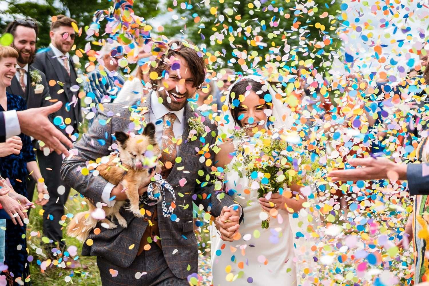 A bride and groom are overwhelmed with bright paper confetti as they leave the church after their ceremony. The groom is holding a small dog and everyone is smiling. The throwing of confetti si the crescendo of our view on how do wedding ceremonies work? 
