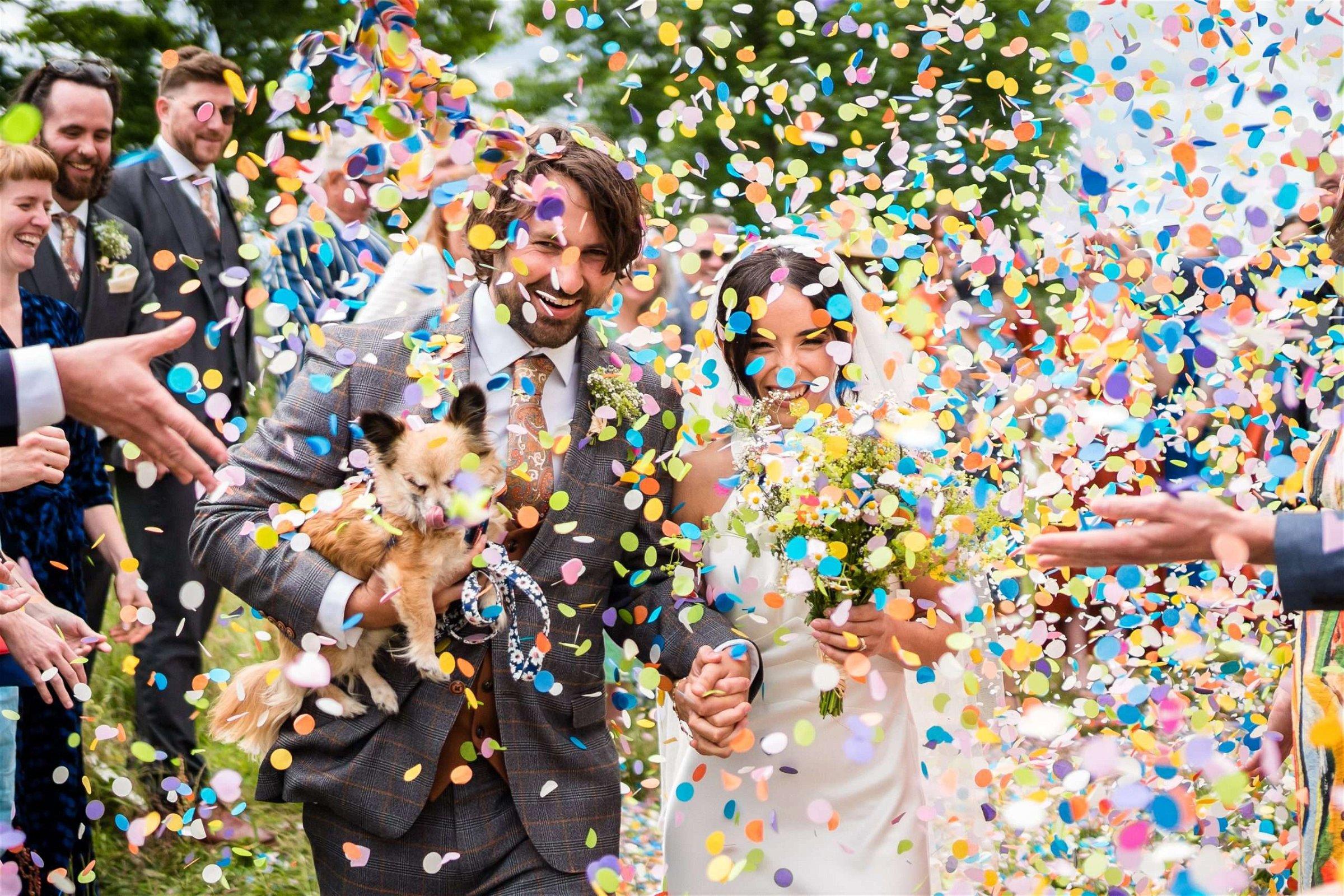 A bride and groom are overwhelmed with bright paper confetti as they leave the church after their ceremony. The groom is holding a small dog and everyone is smiling.