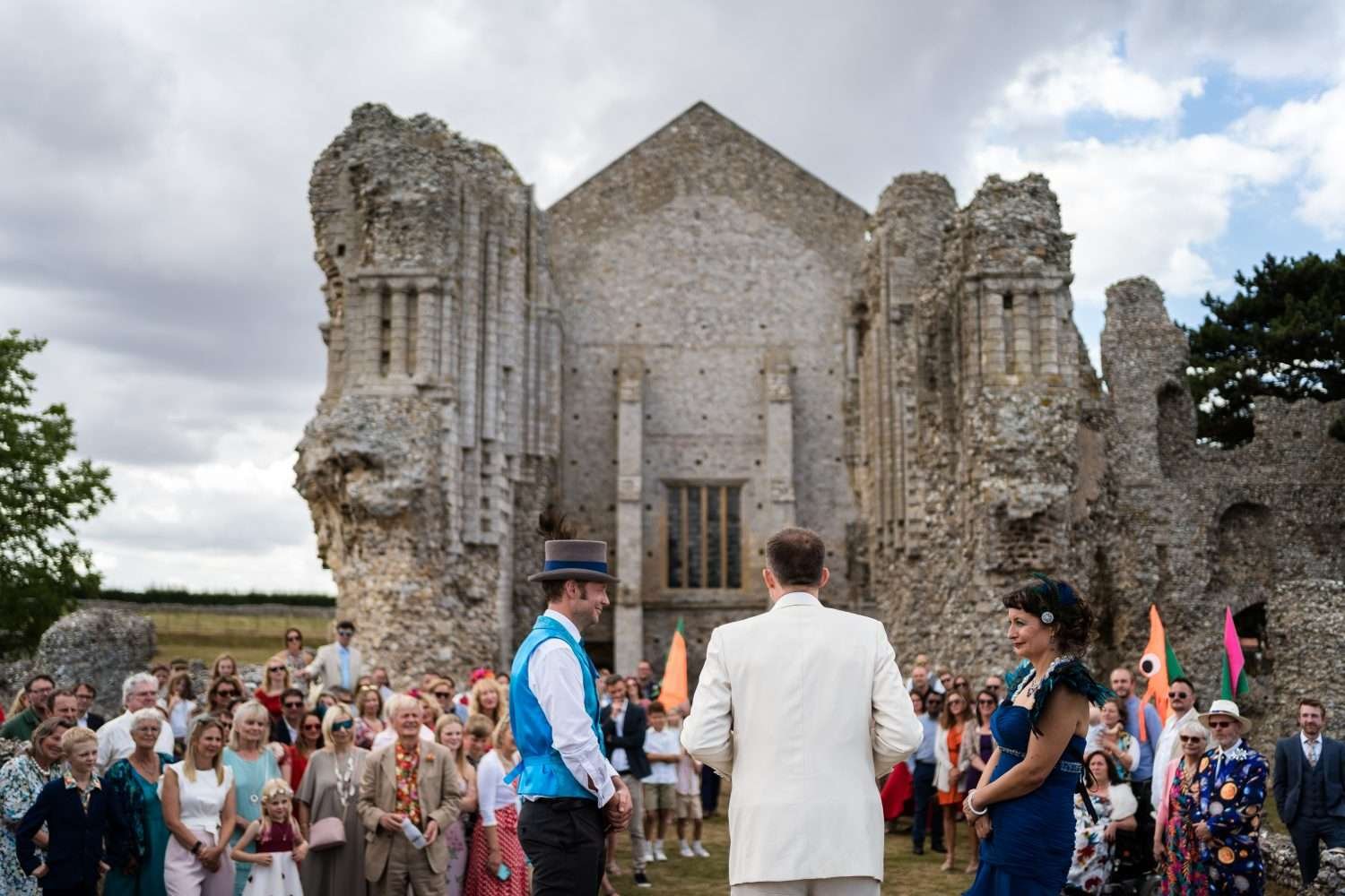 A couple celebrate their marriage at Binham Priory in Norfolk. The celecrant has his back to us as he reads to the family and friends of the family who we see in soft focus behind the couple and with the priory building and ruins set out as the background. 
