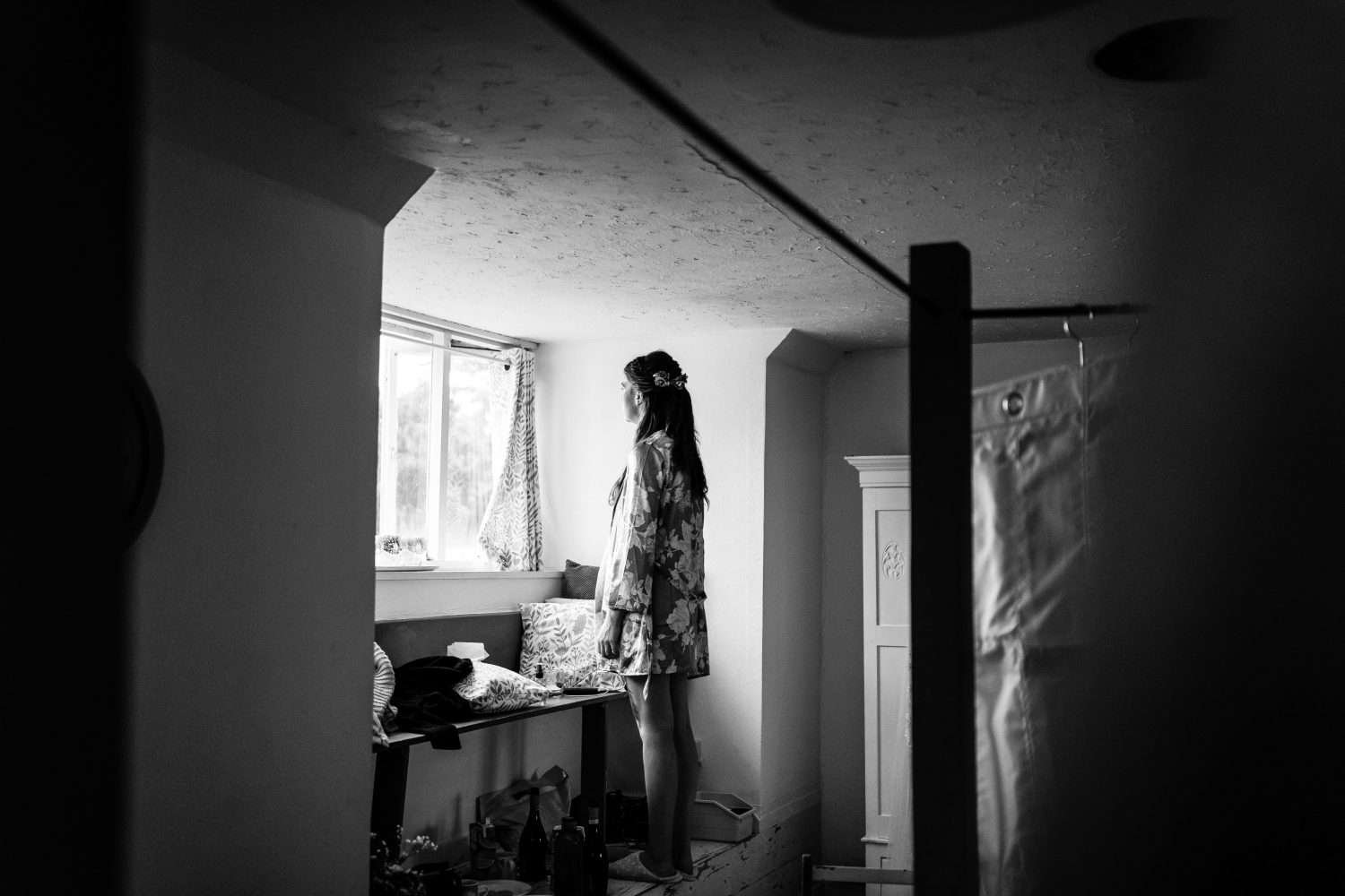A bridesmaid stands in her dressing gown whilst getting ready for a Suffolk village hall wedding. She is looking out of an upstairs window and is illuminated at her front with he back more shadowed. In the foreground and out of focus is the bridal dress hanging from a four poster bed.

