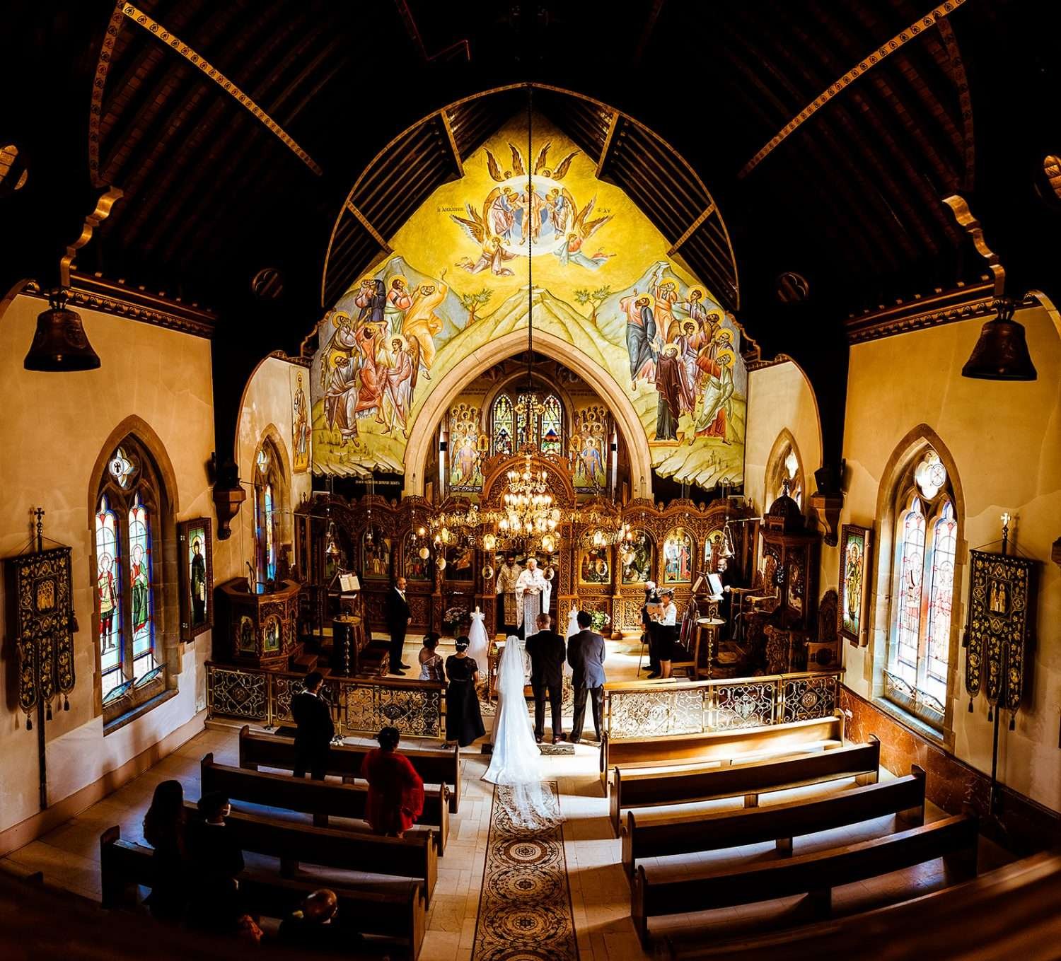 A bride and groom in a Greek Orthodox church during their wedding ceremony. The church is lit by sunlight and the walls and furniture are richly decorated in yellows and gold leaf.