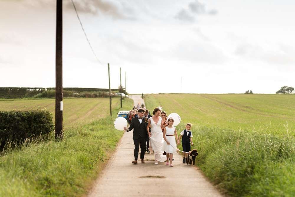 A Fishley Hall wedding scene, The happy couple and all their family and guests parade from the church to fishley hall for the wedding reception. A bridesmaid leads the way holding a basket and with her dog companion. 