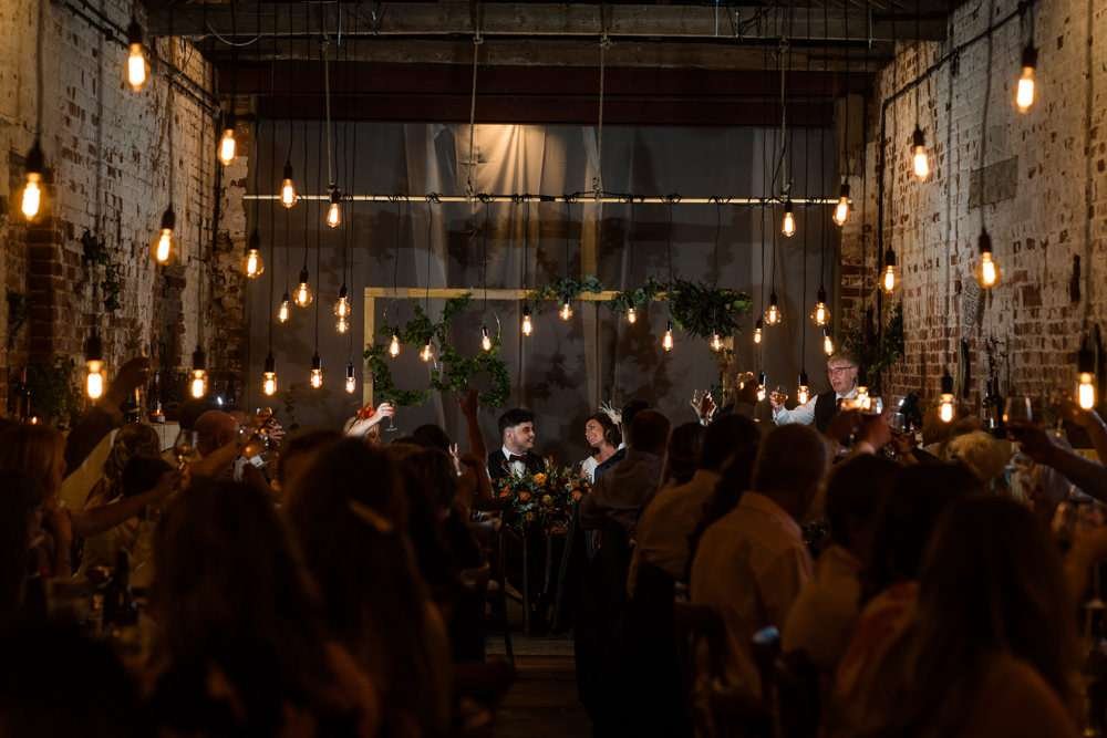 A Bride and Groom are lit by dozens of vintage light pendants in the great barn at their Fishley Hall wedding in Norfolk. They are seated during the wedding breakfast with their friends and family surrounding them.