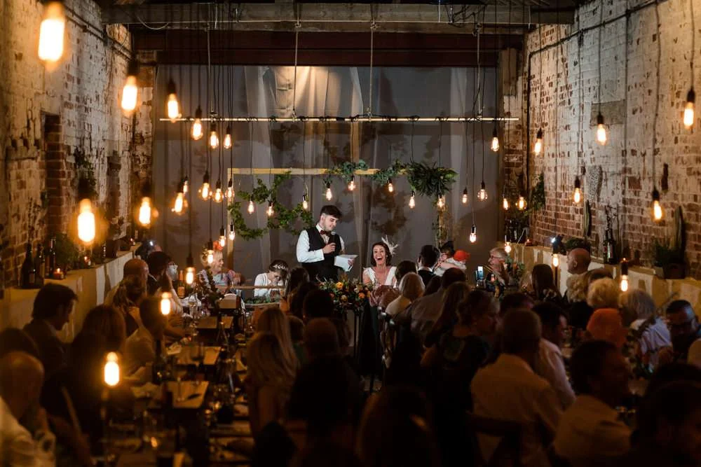A Bride and Groom are lit by dozens of vintage light pendants in the great barn at their Fishley Hall wedding in Norfolk. They are seated during the wedding breakfast with their friends and family surrounding them.