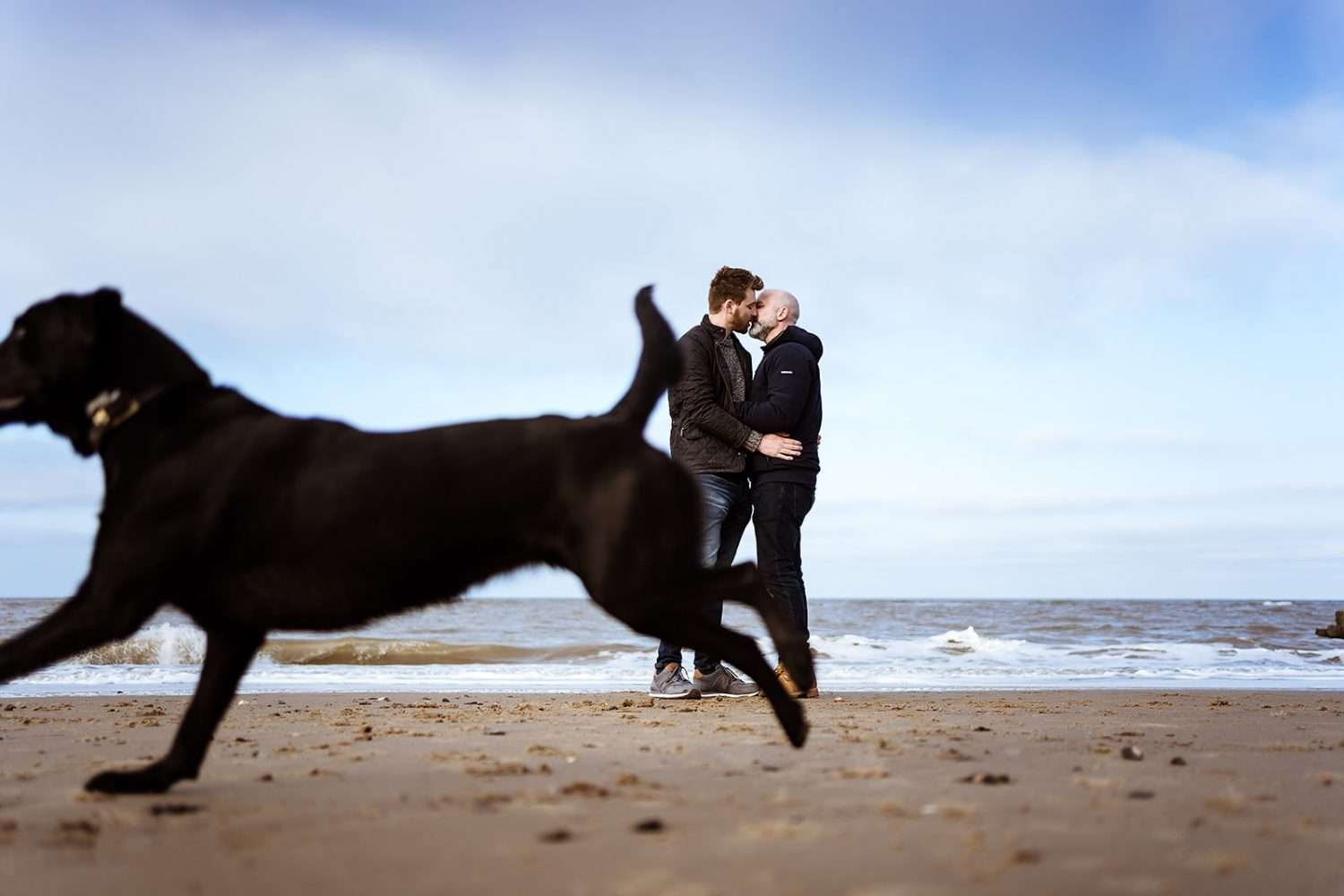 Comfortable natural clothing demonstrating What to wear to your engagement shoot as this same sex couples dog hurtles past them kissing at the beach at cart gap 
