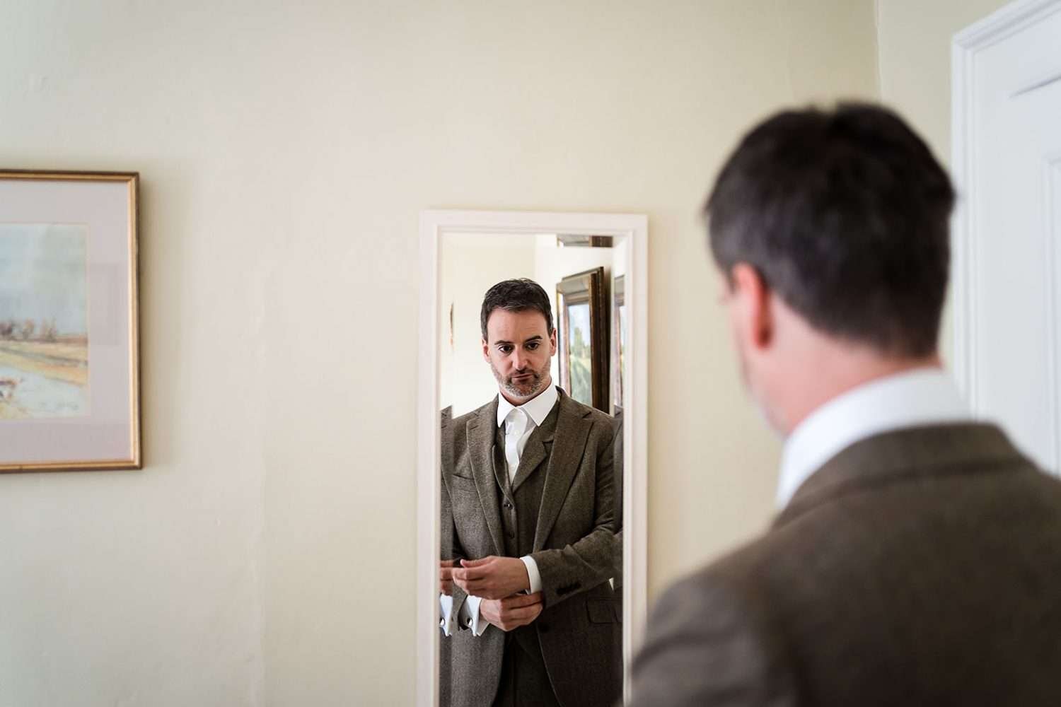 Groom finishes dressing fo rhis bruisyard hall wedding. He's looking in the mirror and adjusting his shirt. 