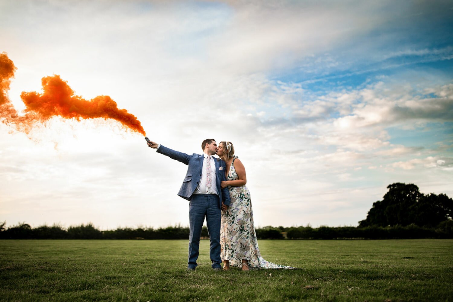 Couple have a wedding portrait in a field in norfolk the groom is holding an orange smoke bomb as they kiss
