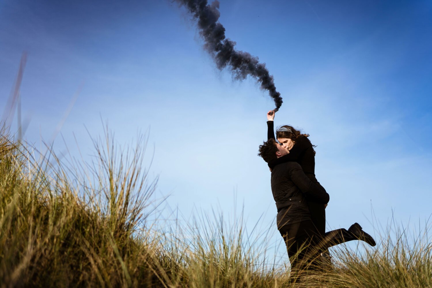 A couple kiss in the dunes at a norfolk beach as the lady holds a black smoke bomb in the air