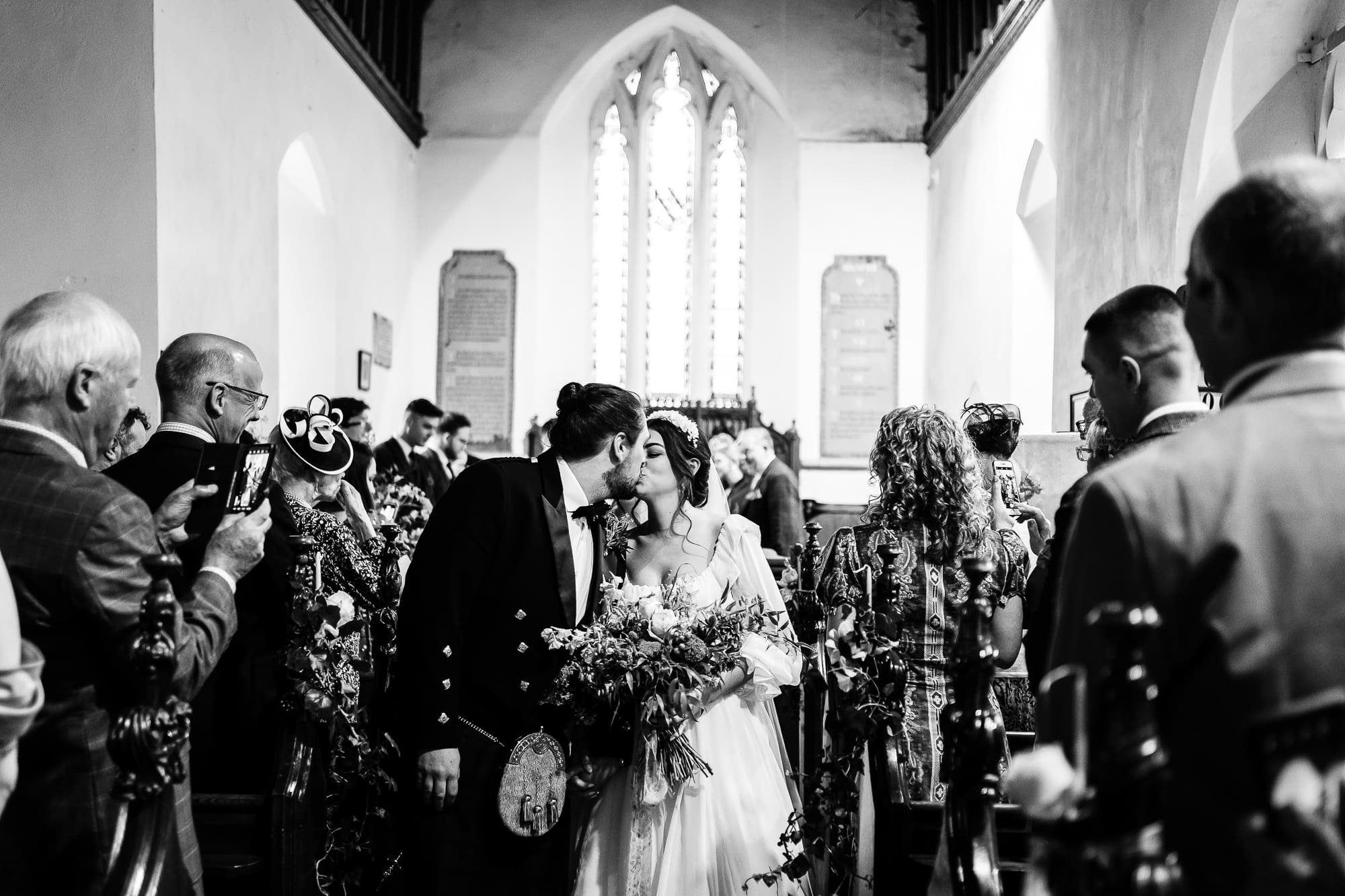 Bride and Groom have stopped down the middle of the aisle in a church to kiss whilst their guests stood in the pews smile, clap and take photos