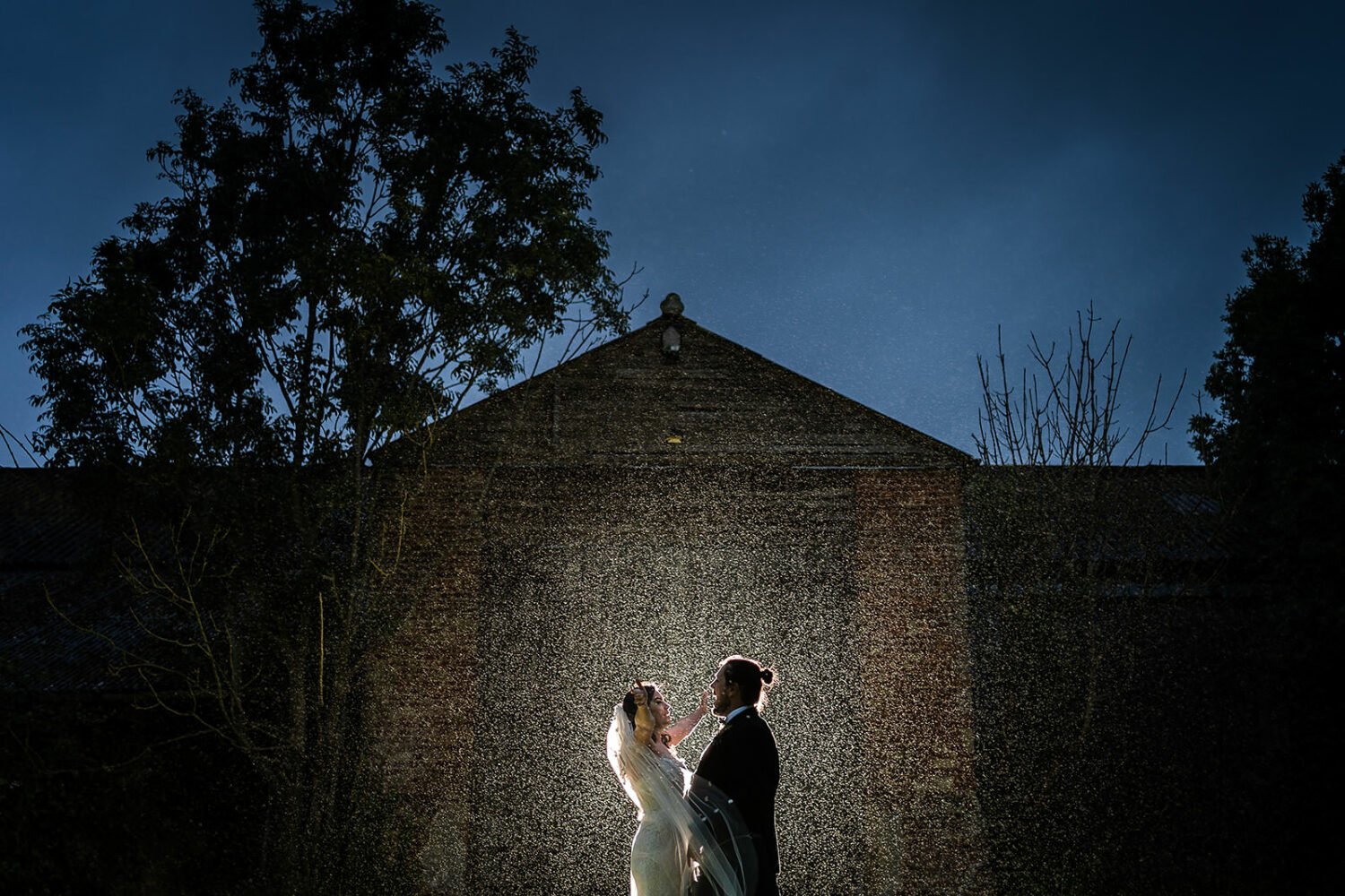 Fishley hall a couple portrait in front of the barn in heavy rain, it is evening and the couple are lit from behind by flash light. The rain drops sparkle.