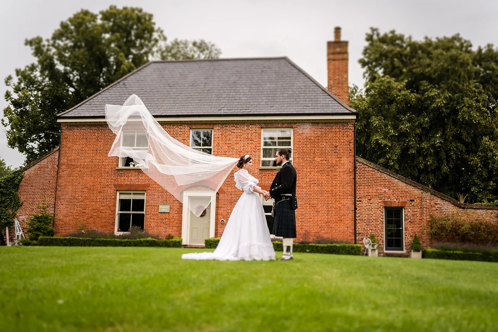 couple hold hands outside the hall at their fishley hall wedding. He is in full traditional Scottish regalia and she has a beautiful dress which has a veil blowing in the wind.
