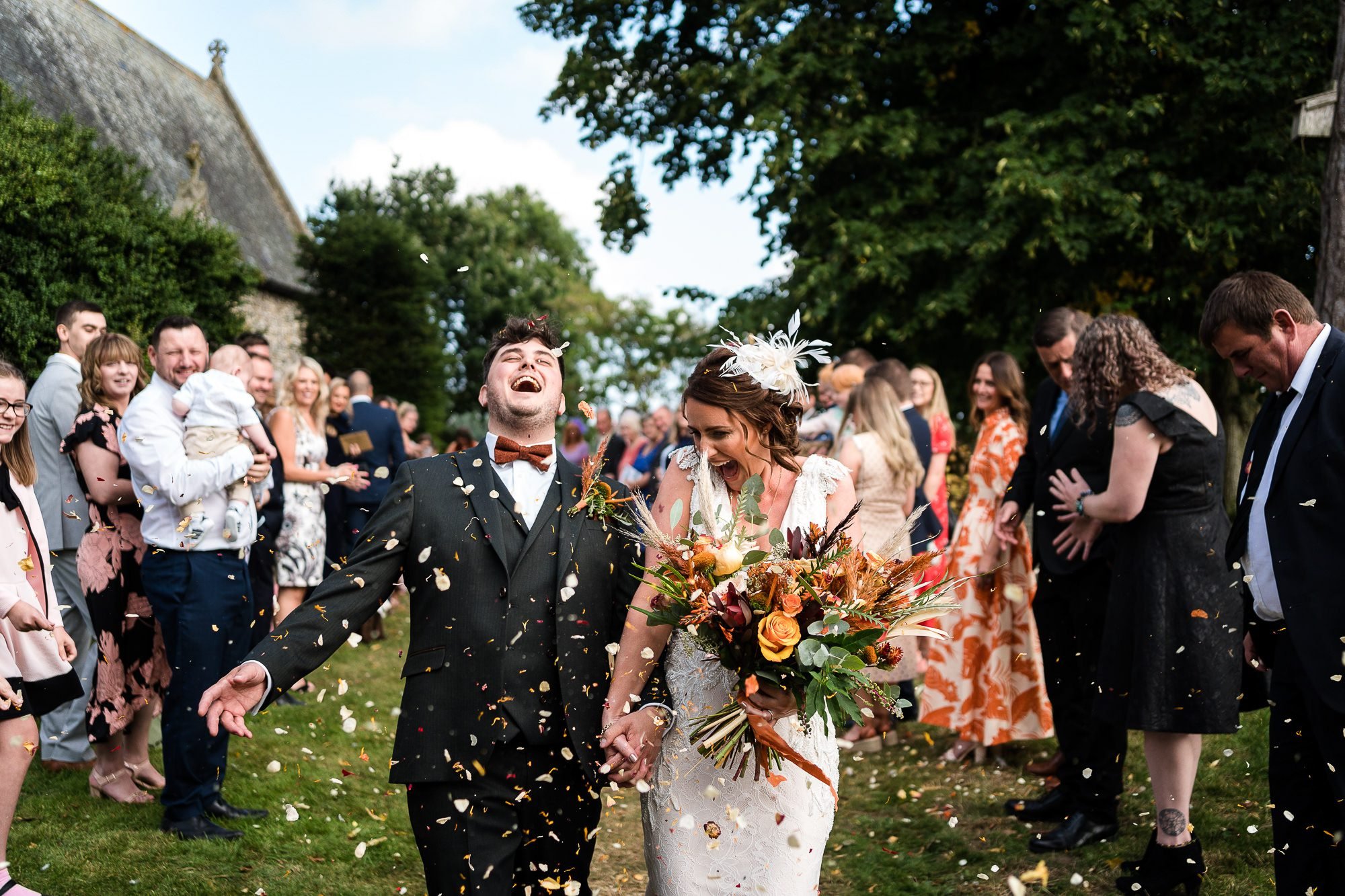 A Bride and Groom laugh and smile as they walk down a confetti line outside the church. They are covered in dried petals which are also in the air