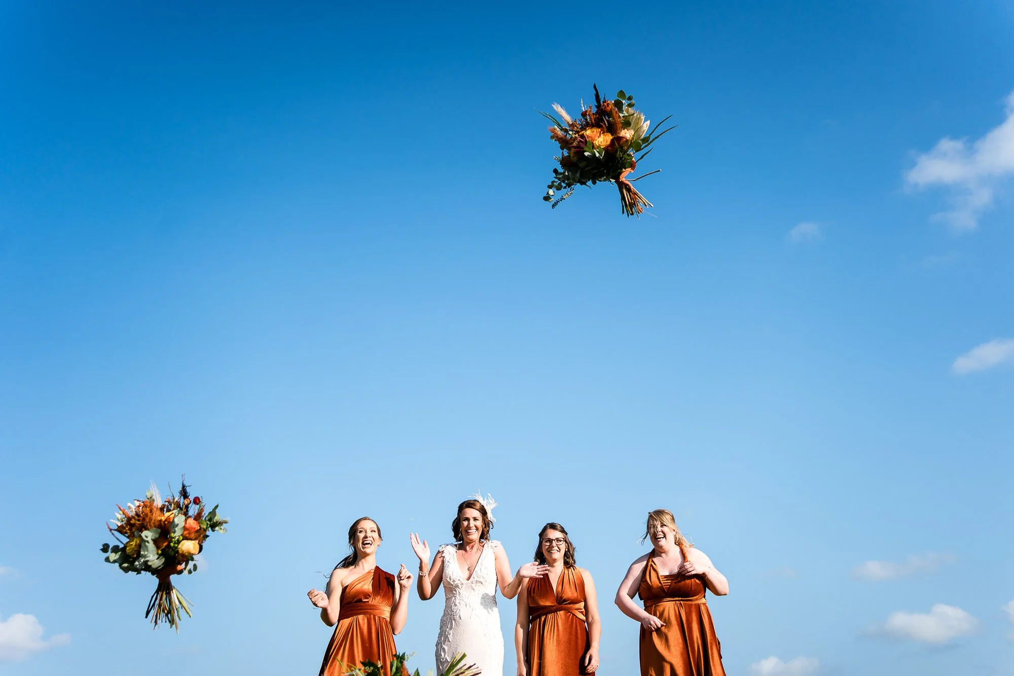 Bride and Bridesmaids stand against blue Norfolk sky and laugh and smile as they have thrown their bouquets in the air