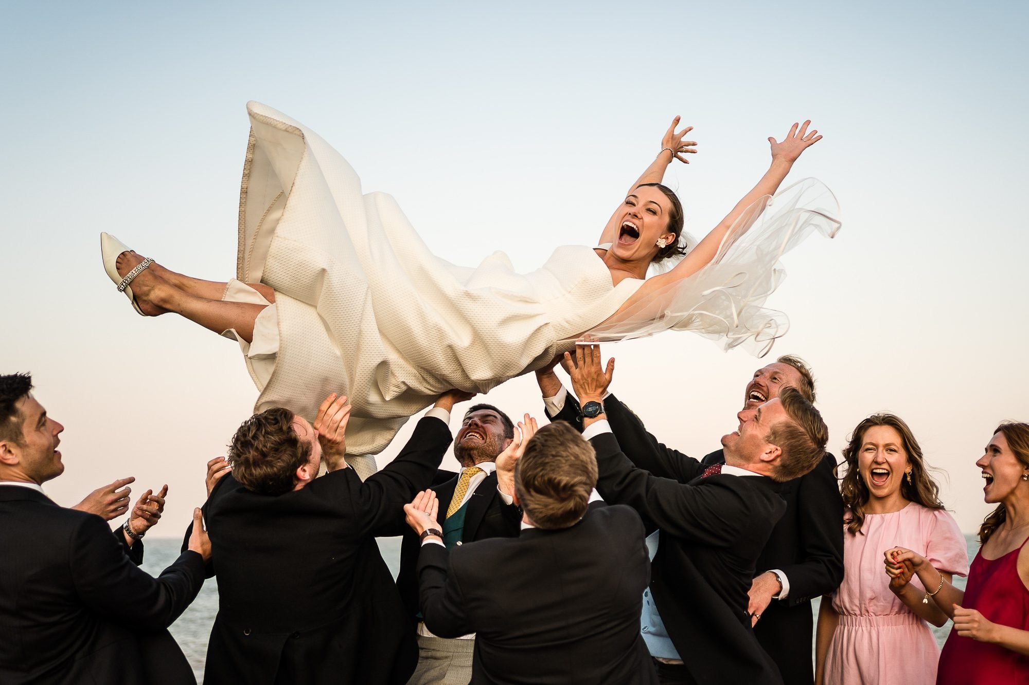 A bride laughs as she is thrown into the air by Groomsmen and her groom who are also laughing. Seaside location