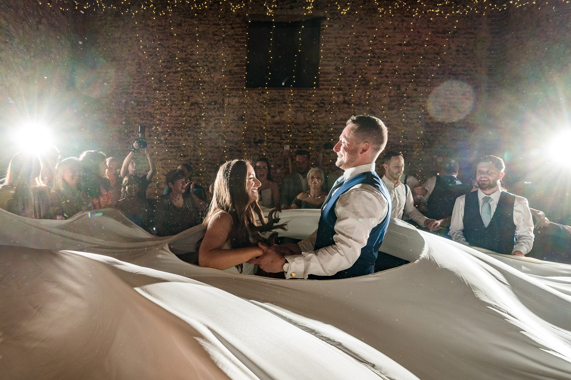 Bride and Groom hold hands and smile as they dance in the middle of a giant sheet as part of Jewish dancing