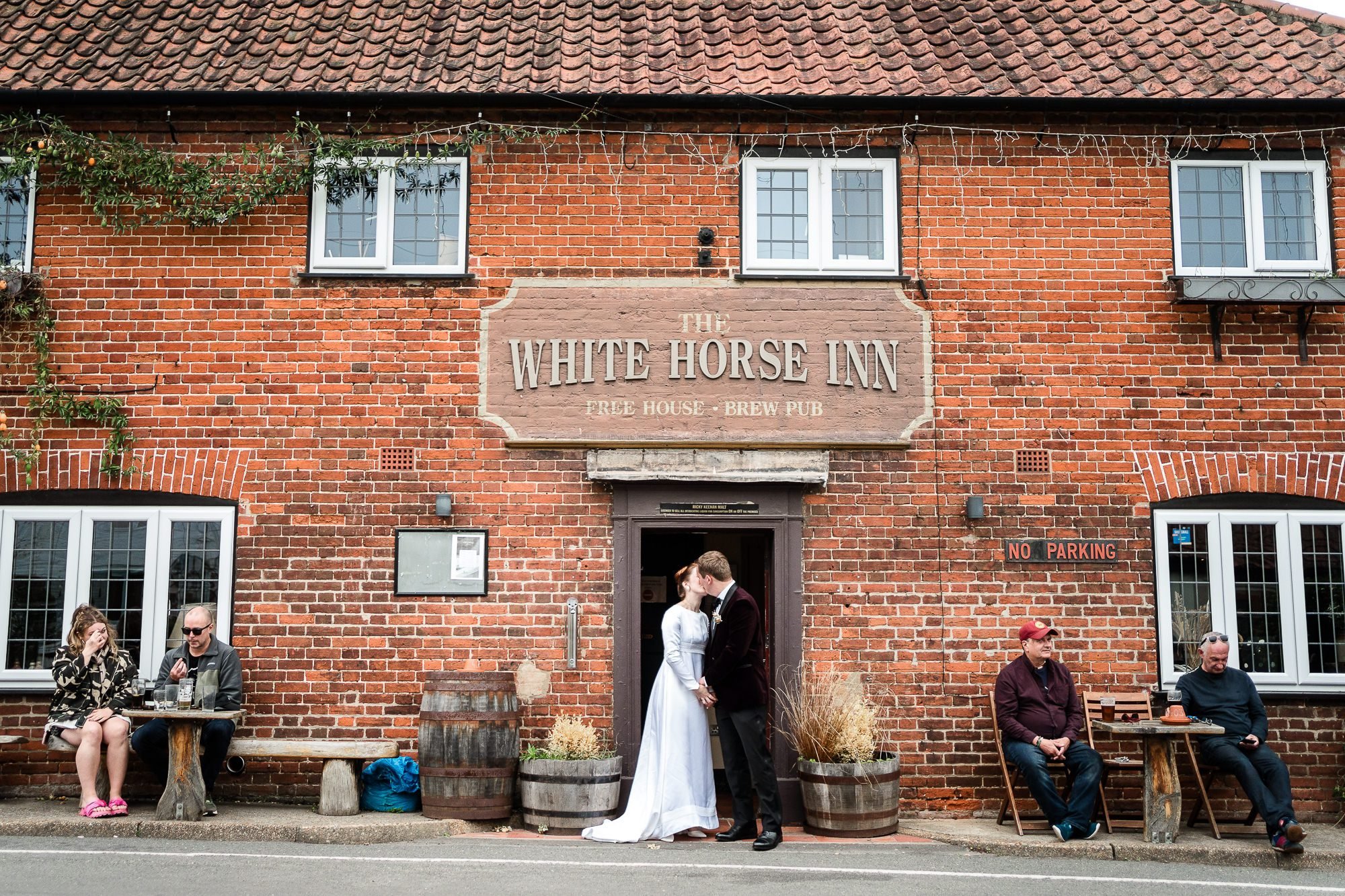 A bride and groom kiss outside The White Horse Inn and Neatishead. There are locals sat around near me enjoying a pint wearing casual clothing